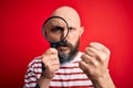 Handsome detective bald man with beard using magnifying glass over red background annoyed and frustrated shouting with anger,