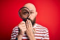 Handsome detective bald man with beard using magnifying glass over red background cover mouth with hand shocked with shame for Royalty Free Stock Photo