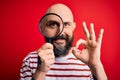 Handsome detective bald man with beard using magnifying glass over red background doing ok sign with fingers, excellent symbol