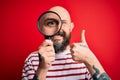 Handsome detective bald man with beard using magnifying glass over red background happy with big smile doing ok sign, thumb up