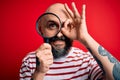 Handsome detective bald man with beard using magnifying glass over red background with happy face smiling doing ok sign with hand