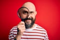 Handsome detective bald man with beard using magnifying glass over red background with a happy face standing and smiling with a