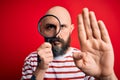 Handsome detective bald man with beard using magnifying glass over red background with open hand doing stop sign with serious and