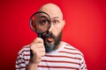 Handsome detective bald man with beard using magnifying glass over red background scared in shock with a surprise face, afraid and Royalty Free Stock Photo