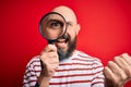 Handsome detective bald man with beard using magnifying glass over red background screaming proud and celebrating victory and