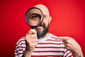 Handsome detective bald man with beard using magnifying glass over red background with surprise face pointing finger to himself