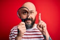 Handsome detective bald man with beard using magnifying glass over red background surprised with an idea or question pointing
