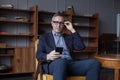 Handsome elderly grey haired man in glasses looking on his mobile phone, sitting on chair in office Royalty Free Stock Photo