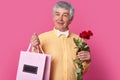 Handsome elegant man wears formal outfit, holds giftbag and bouquet of flowers, going to make present to his wife, being in high