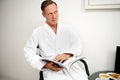 Handsome European middle aged business man wearing white terry bathrobe, sitting on an armchair and reading magazine while resting Royalty Free Stock Photo