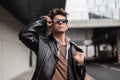 Handsome european young hipster man with trendy hairstyle in fashion leather black jacket straightens stylish sunglasses on a Royalty Free Stock Photo