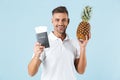 Handsome excited happy adult man posing isolated over blue wall background holding passport and pineapple Royalty Free Stock Photo