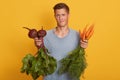 Handsome fairhaired man holding fresh vegetables in hands, young raw foodist decides what to eat beets or carrots isolated over