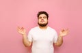 Handsome fat man with curly hair and beard meditates on a pink background with closed eyes wearing a white T-shirt. Authentic