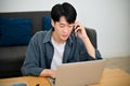 Handsome and focused Asian man talking on the phone while using laptop in living room Royalty Free Stock Photo