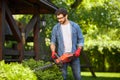 Handsome gardener trimming overgrown conifer bush by electric hedge clippers in summer.