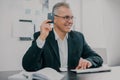 Handsome gray-haired businessman in elegant suit sits in office holding bank card in hand looking excited while working