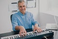 Handsome gray-haired smiling man with earphones sits in music studio playing keyboard piano enjoying music looking happy, music
