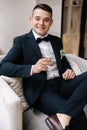 Handsome groom in dark blue suit sitting on chair with glass of whiskey. Groom& x27;s morning. Wedding day