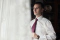 Handsome groom preparing for wedding, putting on tie near window