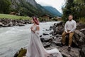 handsome groom sitting on rock and looking at beautiful tender bride standing near mountain river
