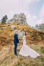 Handsome groom in stylish blue suit holding hands of his charming white dressed bride on idyllic rocky landscape as