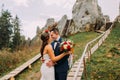 Handsome groom in stylish blue suit kissing his white dressed bride holding bouquet of roses on majestic mountain Royalty Free Stock Photo