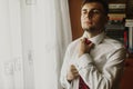 Handsome groom in stylish white shirt putting on elegant red tie