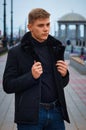 Handsome guy in a black jacket with a fur collar against the background of the city embankment with a rotunda in blur. Royalty Free Stock Photo