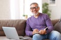 Handsome happy senior man in glasses working remotely while sitting on sofa with laptop computer Royalty Free Stock Photo