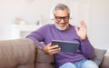 Handsome happy senior man waving by hand while talking online on digital tablet with family Royalty Free Stock Photo