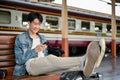 A happy Asian male backpacker using his smartphone on a bench while waiting for his train Royalty Free Stock Photo