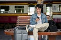 A happy Asian male backpacker using his phone while waiting for his train on a bench at the platform Royalty Free Stock Photo