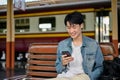 A happy Asian male backpacker using his smartphone while waiting for his train on a bench Royalty Free Stock Photo