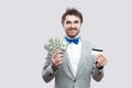 Handsome happy young bearded man in gray suit and blue bow tie standing and holding bank credit card and many cash dollars with