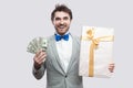 Handsome happy young bearded man in gray suit and blue bow tie standing and holding gift box with yellow bow and many cash dollars Royalty Free Stock Photo