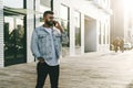 Handsome hipster businessman with beard, in denim jacket and trendy glasses walks around city and calls on mobile phone. Royalty Free Stock Photo