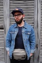 Handsome hipster guy in a cap and glasses with a beard in a blue denim jacket and a fanny pack