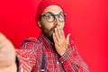 Handsome hipster man with beard taking a selfie photo with smartphone covering mouth with hand, shocked and afraid for mistake