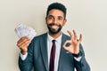 Handsome hispanic business man with beard holding swedish krona banknotes doing ok sign with fingers, smiling friendly gesturing Royalty Free Stock Photo