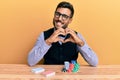 Handsome hispanic croupier man sitting on the table with poker chips and cards smiling in love showing heart symbol and shape with