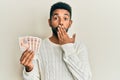 Handsome hispanic man with beard holding 10 united kingdom pounds banknotes covering mouth with hand, shocked and afraid for