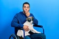 Handsome hispanic man with beard sitting on wheelchair with puppy dog covering mouth with hand, shocked and afraid for mistake Royalty Free Stock Photo
