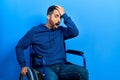 Handsome hispanic man with beard sitting on wheelchair surprised with hand on head for mistake, remember error