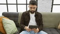 Handsome hispanic man with beard using tablet on couch in modern apartment living room Royalty Free Stock Photo
