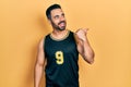 Handsome hispanic man with beard wearing basketball t shirt pointing thumb up to the side smiling happy with open mouth Royalty Free Stock Photo