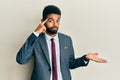 Handsome hispanic man with beard wearing business suit and tie confused and annoyed with open palm showing copy space and pointing Royalty Free Stock Photo