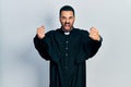 Handsome hispanic man with beard wearing catholic priest robe angry and mad raising fists frustrated and furious while shouting