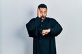 Handsome hispanic man with beard wearing catholic priest robe looking at the watch time worried, afraid of getting late Royalty Free Stock Photo