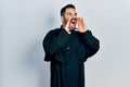 Handsome hispanic man with beard wearing catholic priest robe shouting angry out loud with hands over mouth Royalty Free Stock Photo